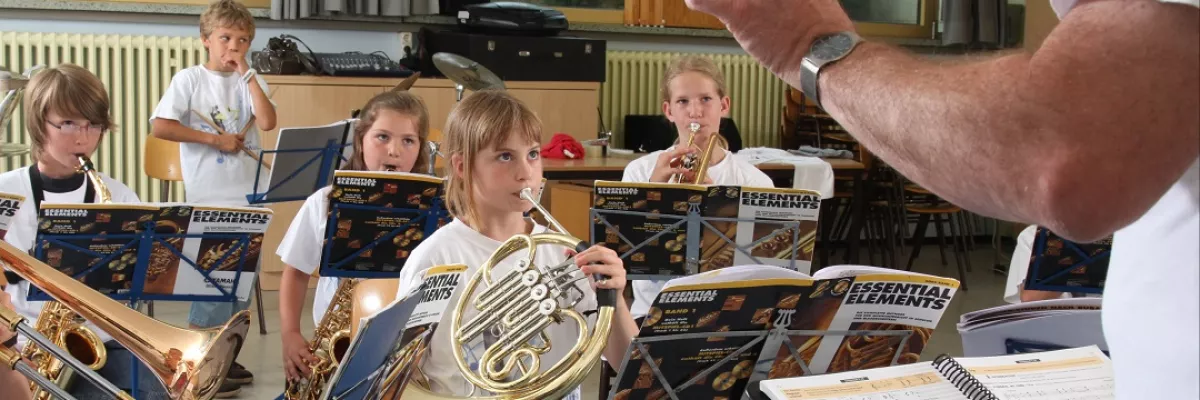 Kinder musizieren gemeinsam und werden von einem Mann dirigiert. 